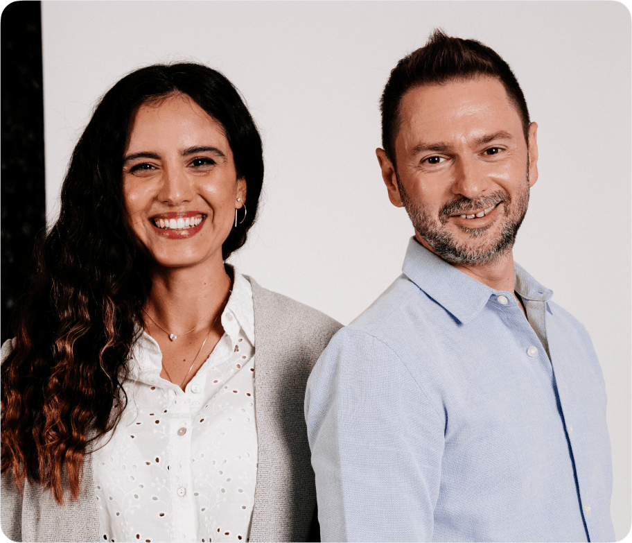 Smiling woman with long curly hair and man with short hair and beard standing side by side against a plain background.