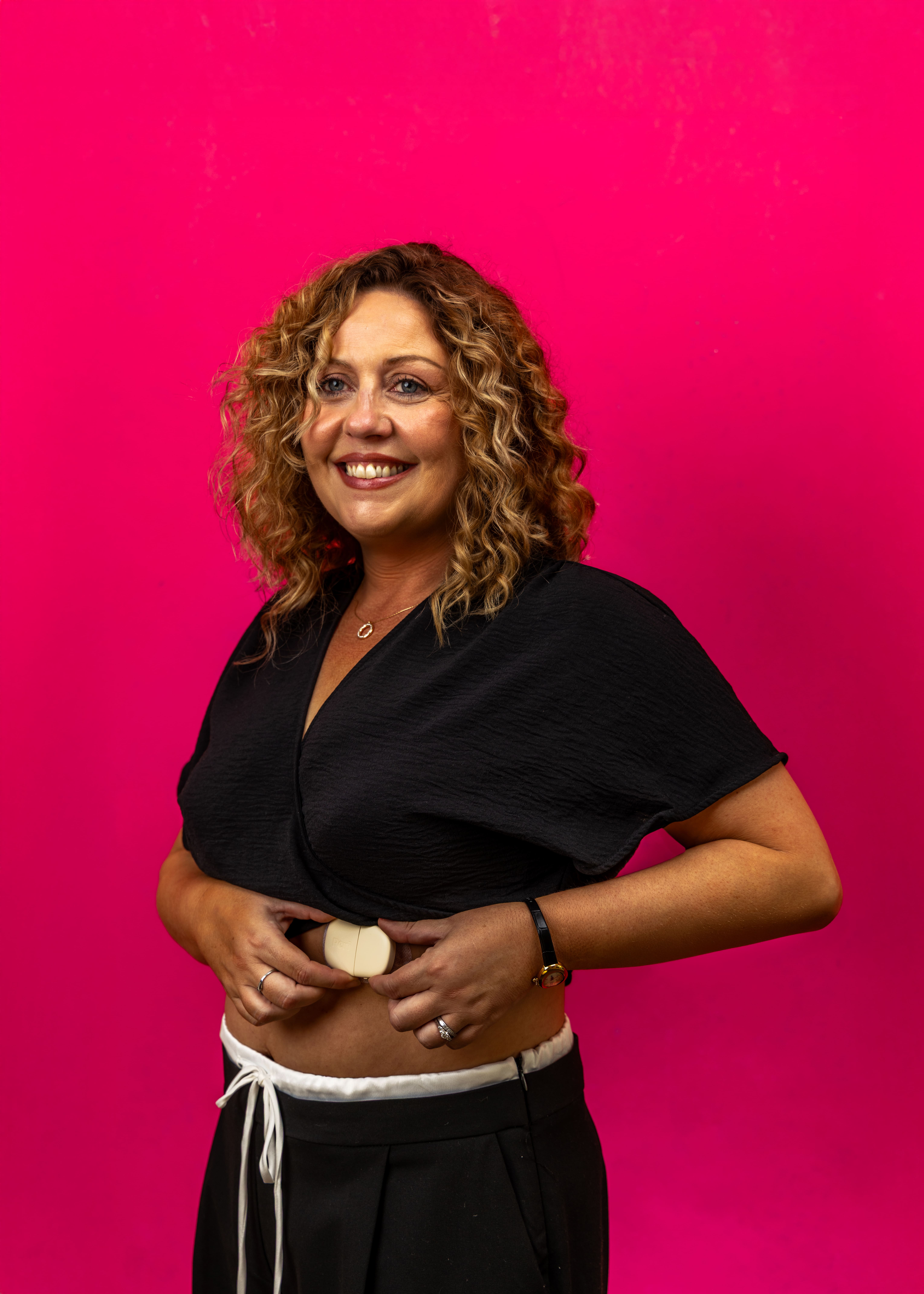 Smiling woman with curly hair adjusting a beige device on her exposed midriff against a bright pink background.