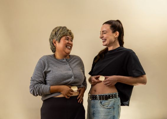 Two women laughing and comparing their stomachs with round objects held at their waists against a neutral background.