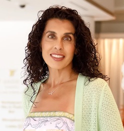 Smiling woman with curly dark hair wearing a light green cardigan and patterned top indoors.