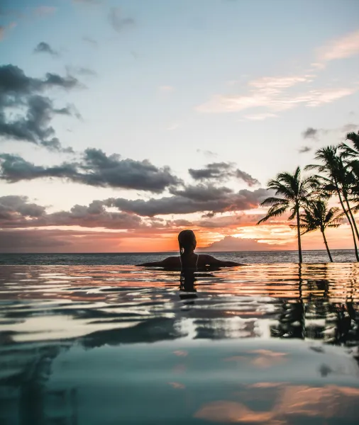 Person with braided hair leaning on edge of infinity pool at sunset with palm trees and ocean in background.