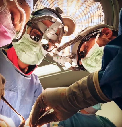 Surgeons wearing masks, protective eyewear, and headlamps performing surgery under bright operating room lights.