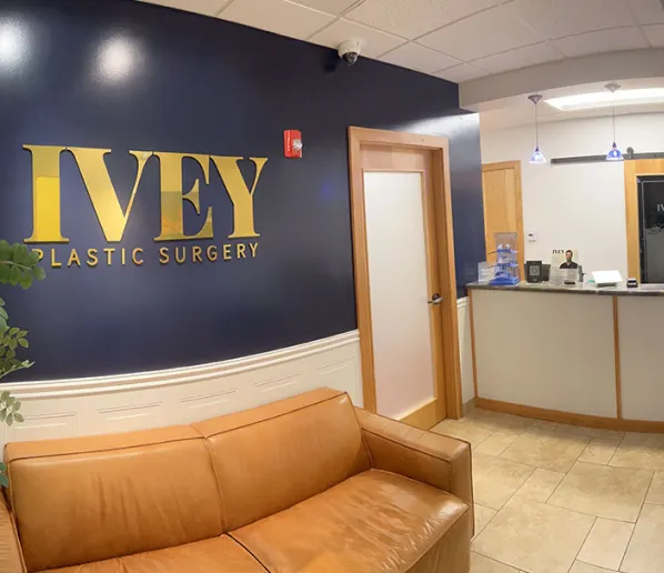 Reception area of IVEY Plastic Surgery with a brown leather couch, navy blue wall with gold lettering, wooden door, and a front desk with a receptionist.
