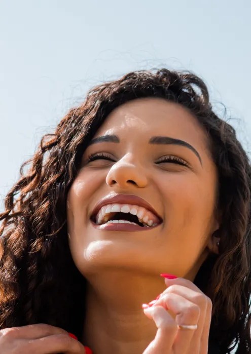Close-up of a young woman with curly hair smiling and looking joyful against a clear sky background.