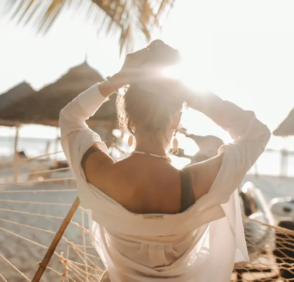 Woman sitting on a hammock at the beach with the sun shining behind her, wearing a white shirt and pearl necklace.