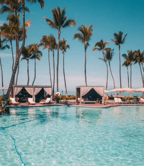 Resort pool with clear blue water, palm trees, and cabanas under a bright sky.