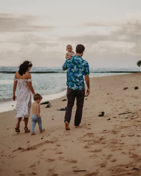 Family of four walking barefoot on a sandy beach near the ocean under a cloudy sky.
