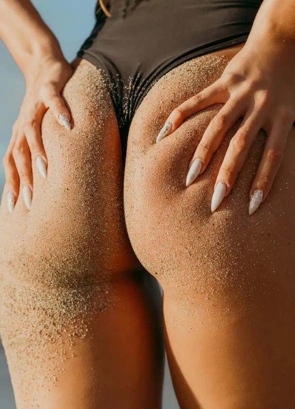 Close-up of a person’s sand-covered thighs and hands with long manicured nails wearing black swimwear.