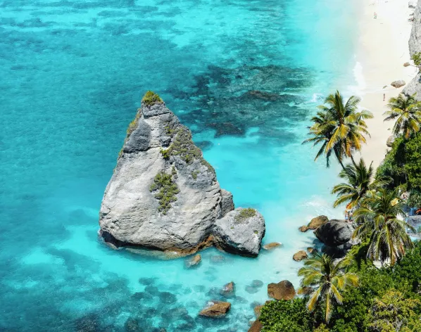 Large rocky formation surrounded by clear turquoise water near a sandy beach lined with palm trees.
