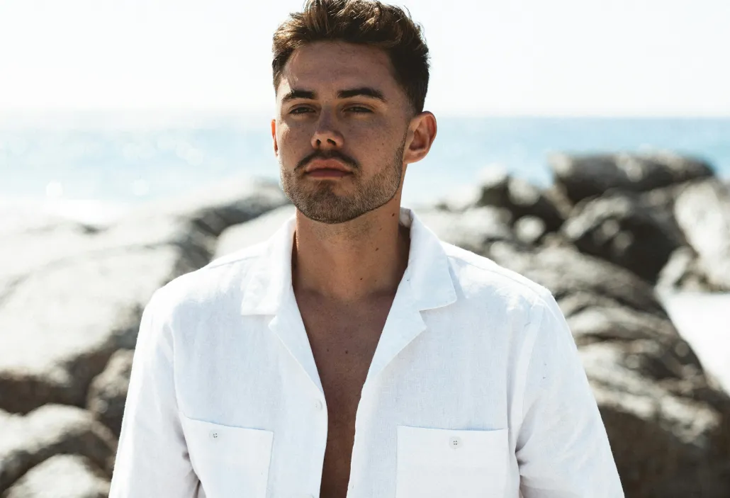 Young man with short dark hair and facial hair wearing an unbuttoned white shirt standing outdoors near rocks and the sea.