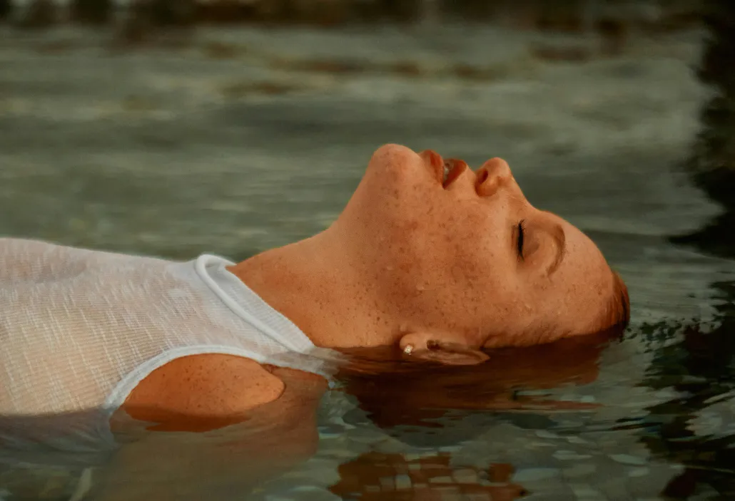 Woman with closed eyes and freckles floating on water wearing a white sleeveless top.