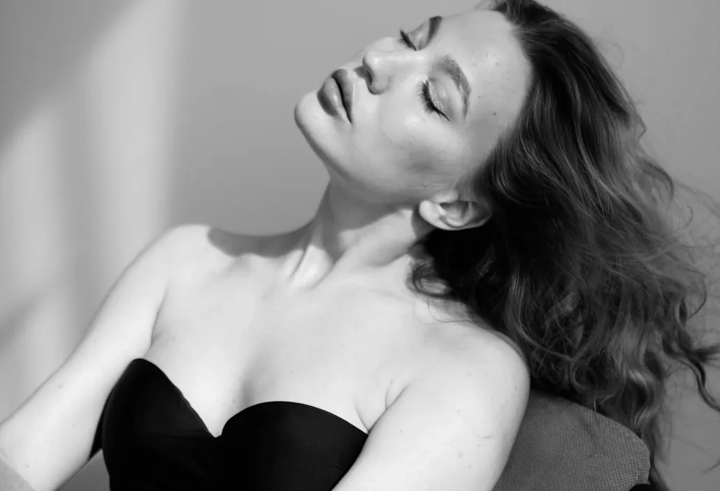 Black and white photo of a woman with long wavy hair leaning back with closed eyes and wearing a strapless top.