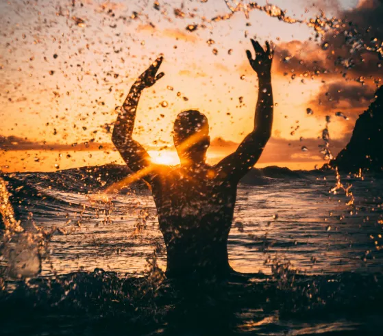 Silhouette of a person splashing water with raised arms at sunset over the ocean.