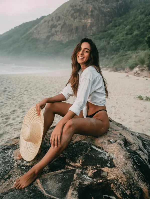 Smiling woman with long hair wearing a white shirt and black bikini bottoms sitting barefoot on a rock at a sandy beach with a straw hat in hand and mountains in the background.