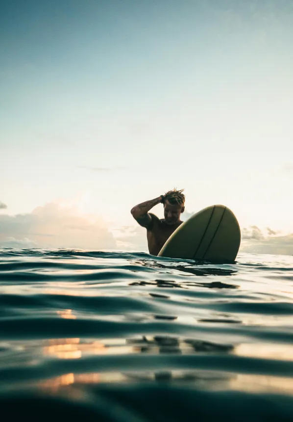 Man standing in the ocean holding a surfboard with one hand on his head during sunset.