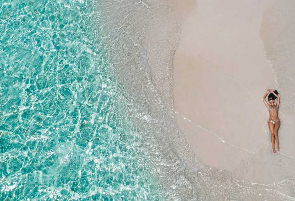 Woman in bikini lying on wet sandy beach beside clear turquoise ocean water.