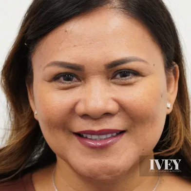 Close-up portrait of a woman with dark hair smiling against a white background, wearing stud earrings and a necklace.