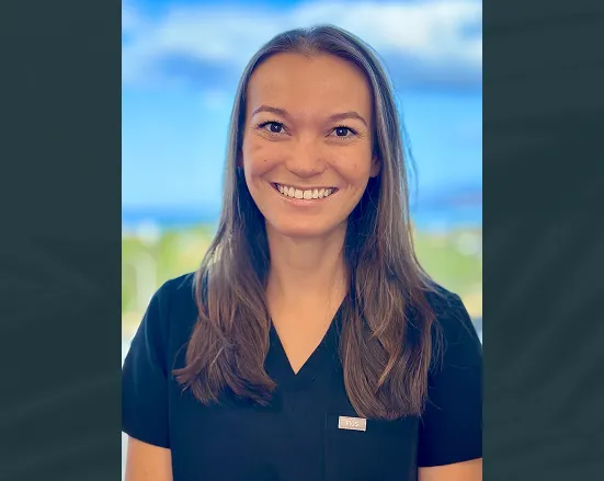 Smiling woman with long brown hair wearing a black scrub top with an FDS pin, blue sky and greenery blurred in the background.