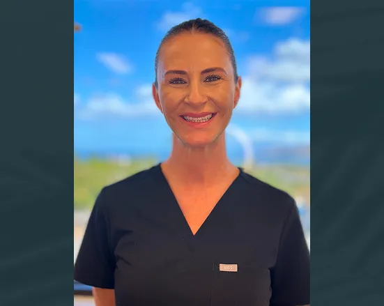 Smiling woman with hair pulled back wearing black medical scrubs against a bright blue sky background.