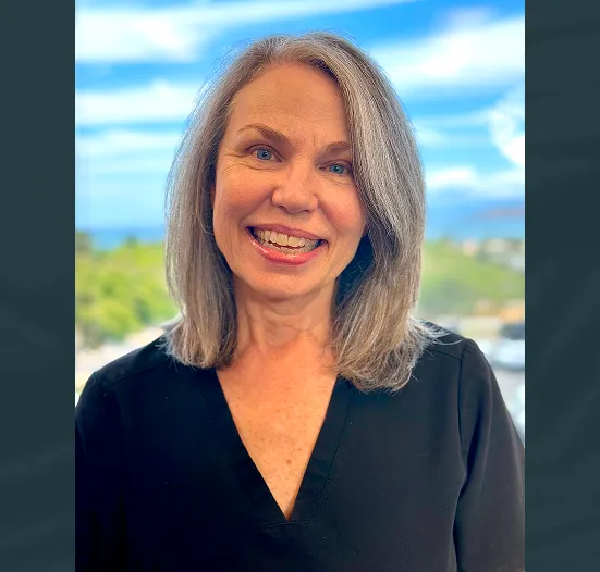 Smiling middle-aged woman with shoulder-length gray hair wearing a black top, standing outdoors with a blurred landscape and blue sky behind her.