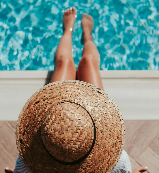 Person wearing a large woven straw hat sitting by the edge of a swimming pool with legs extended into the water.