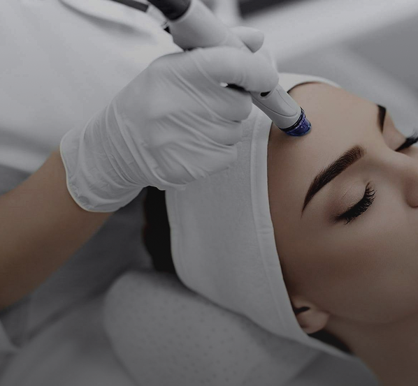 Woman receiving a facial treatment with a handheld device applied to her forehead by a gloved aesthetician.