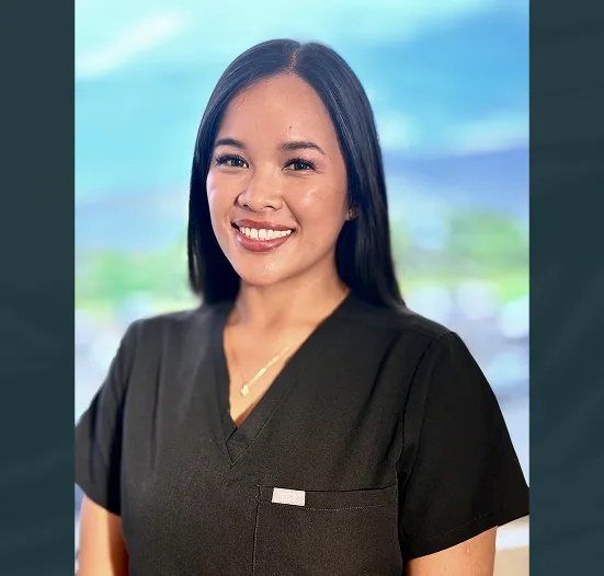 Smiling woman with long dark hair wearing black scrubs standing in front of a blurred window background.