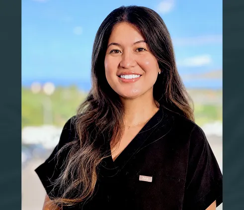 Smiling woman with long dark hair wearing black medical scrubs and a name tag standing outdoors.