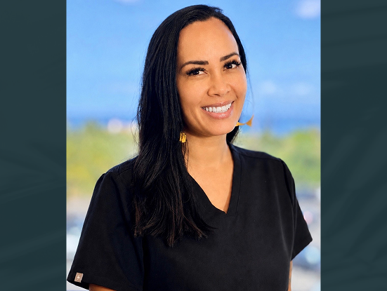 Smiling woman with long dark hair wearing black scrubs standing outdoors with a blurred green background.