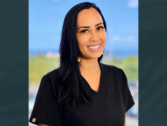 Smiling woman with long dark hair wearing black scrubs standing outdoors with a blurred green background.