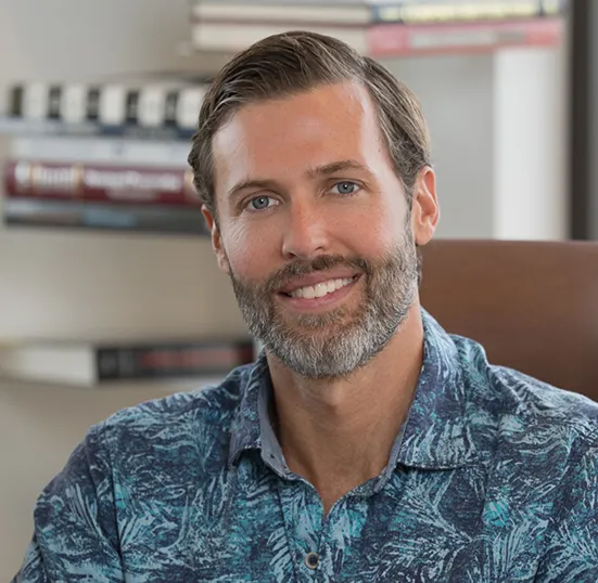 Smiling man with beard wearing a blue patterned shirt sitting in front of blurred bookshelves.