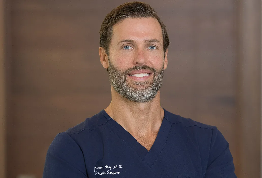 Smiling man with beard in navy blue medical scrubs, with text 'Simon Ivey M.D. Plastic Surgeon' visible.