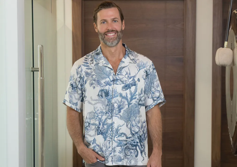Smiling man with beard wearing a patterned short-sleeve shirt standing indoors in front of a wooden door.