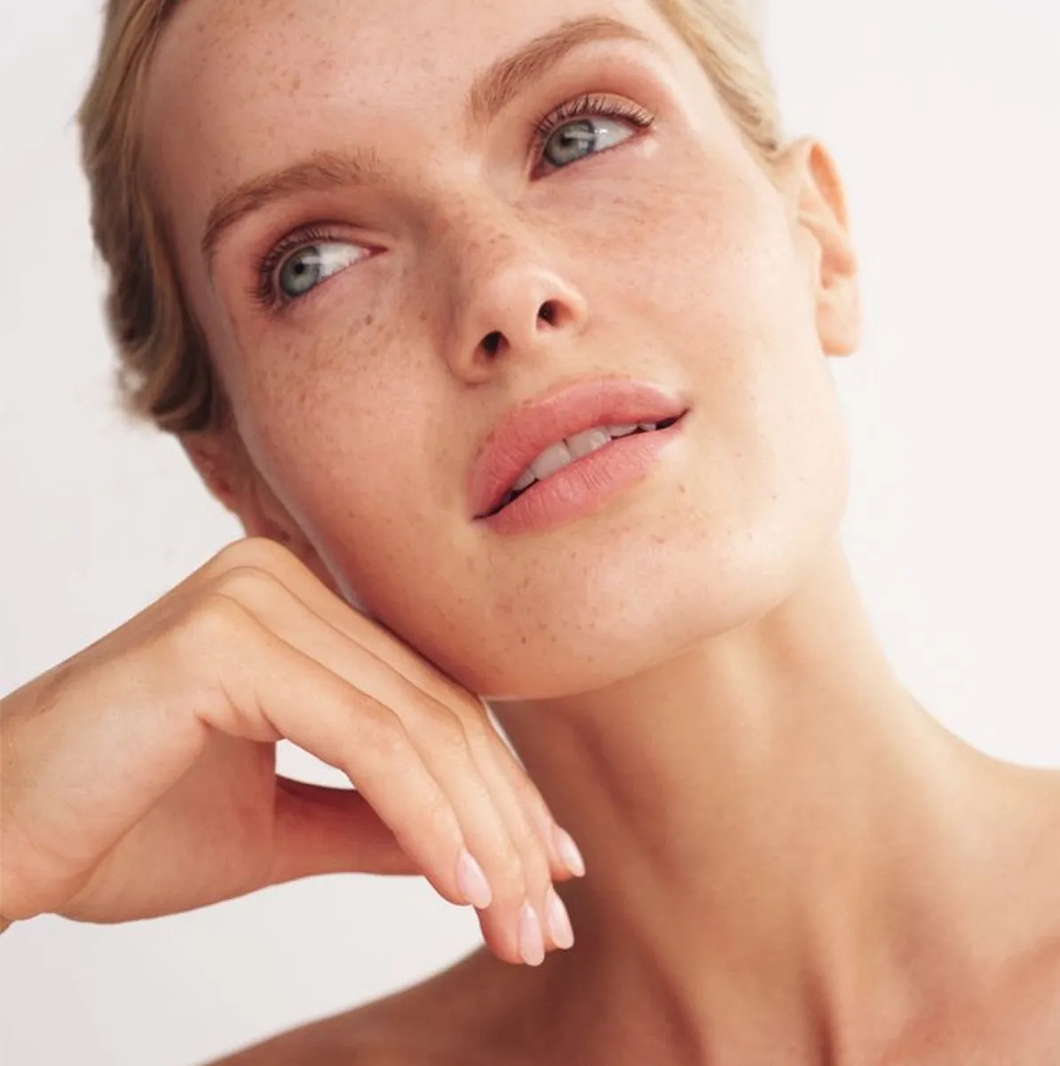Close-up of woman with freckles and light makeup looking thoughtfully to the side with hand near chin.