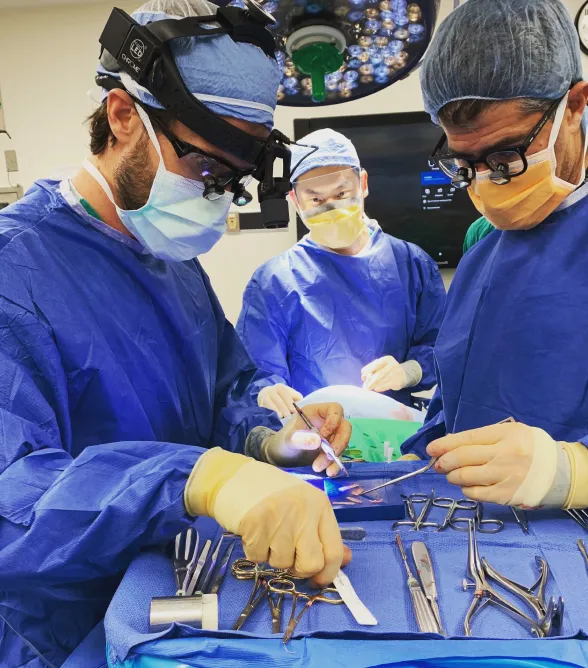 Three surgeons in blue scrubs and masks performing surgery with tools on a sterile drape.