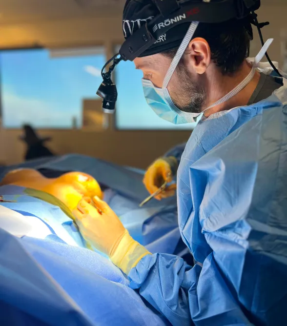 Surgeon in blue scrubs and mask focused on a patient during surgery under bright light.