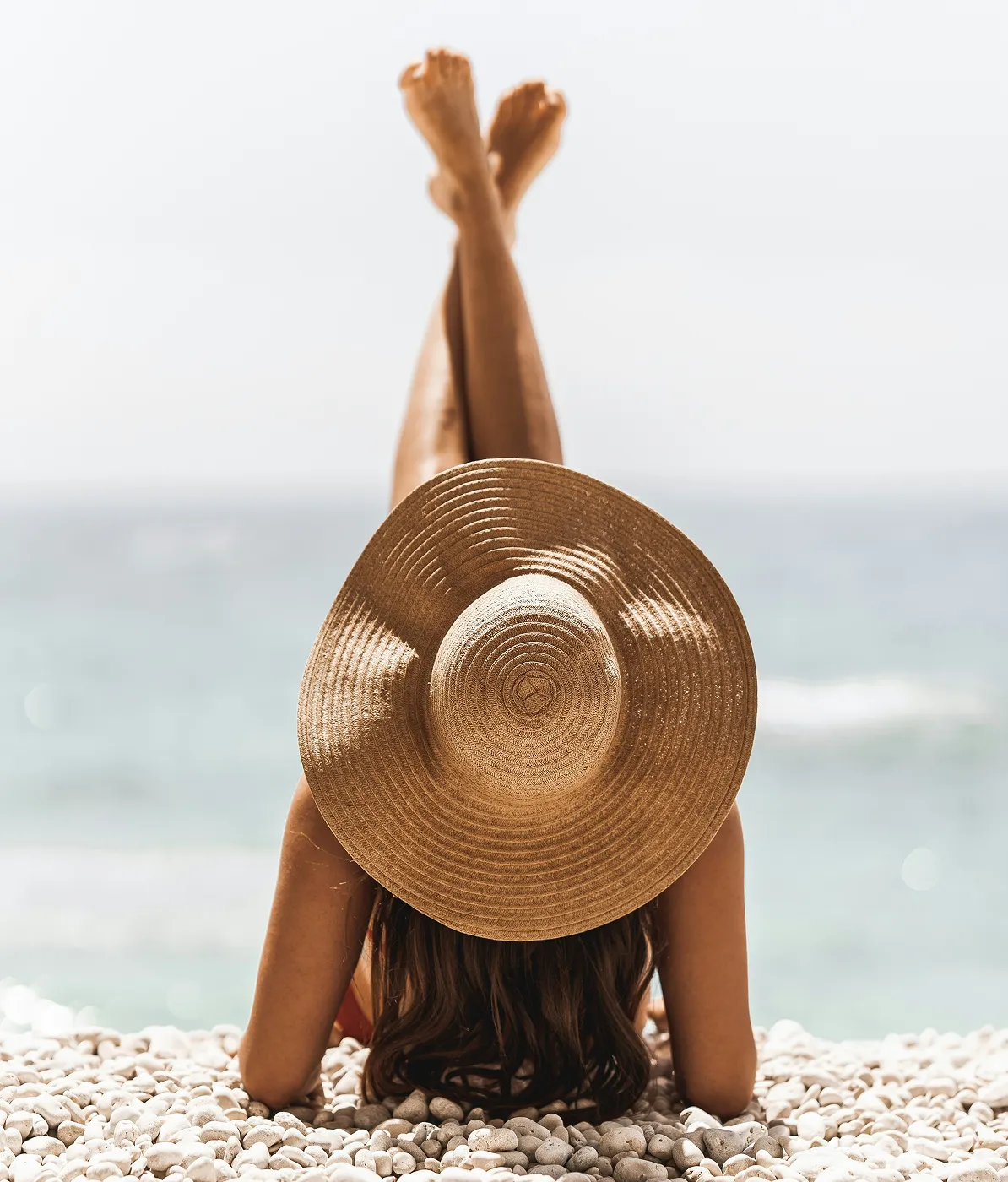 Person lying on pebble beach with legs crossed up, wearing a large sun hat facing the sea.