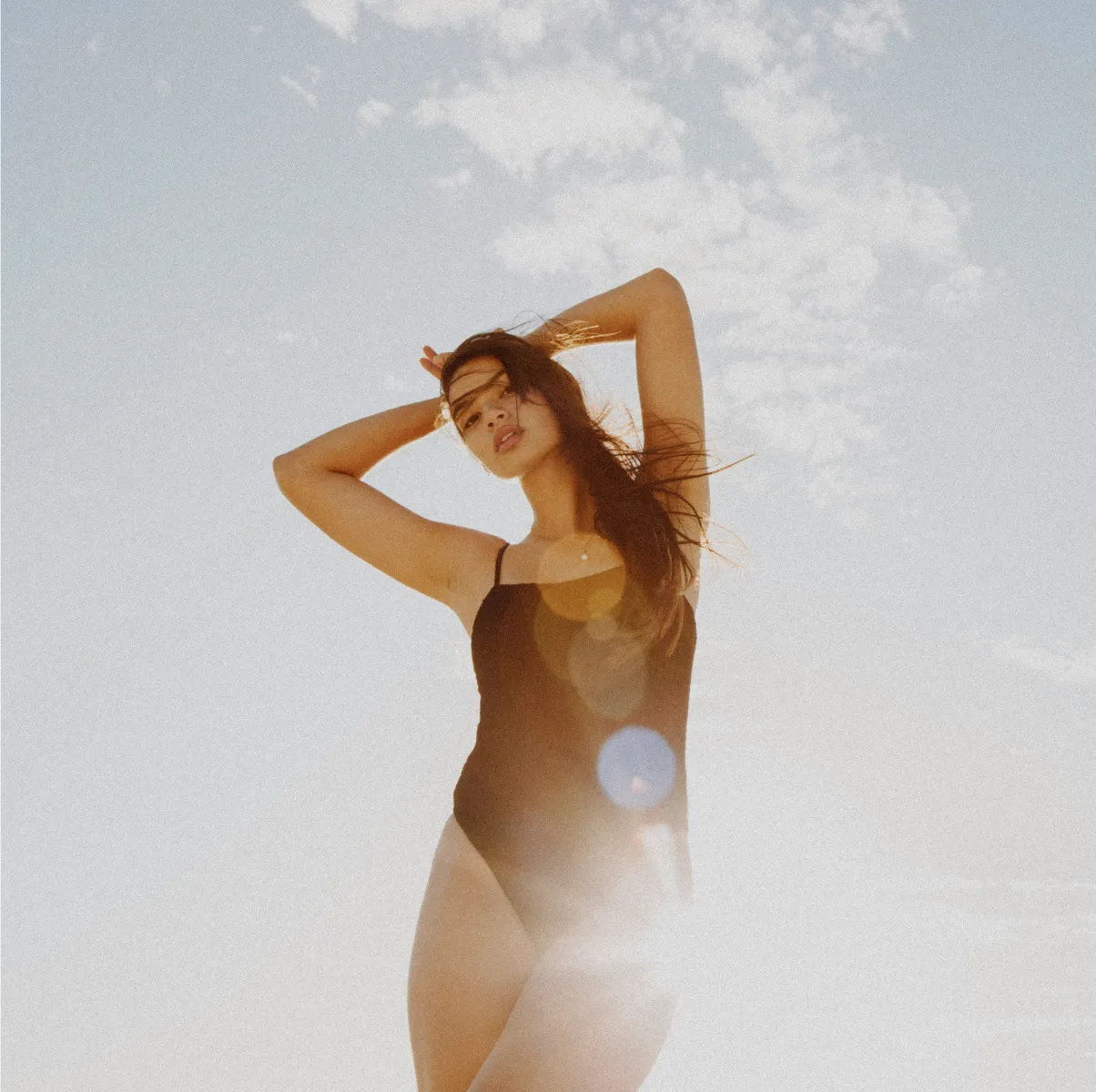 Woman in black swimsuit posing with hands behind head against a bright sky with clouds.