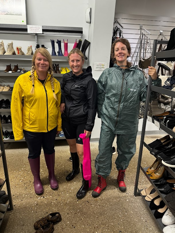 Three women in rain gear and boots stand in a thrift store shoe aisle, laughing at their improvised outfits for the wet weather.