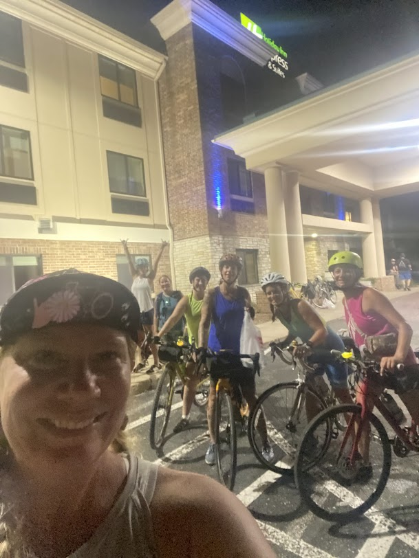 Cyclists pose with big smiles outside a Holiday Inn Express at night, bikes lined up after a long day on the road.
