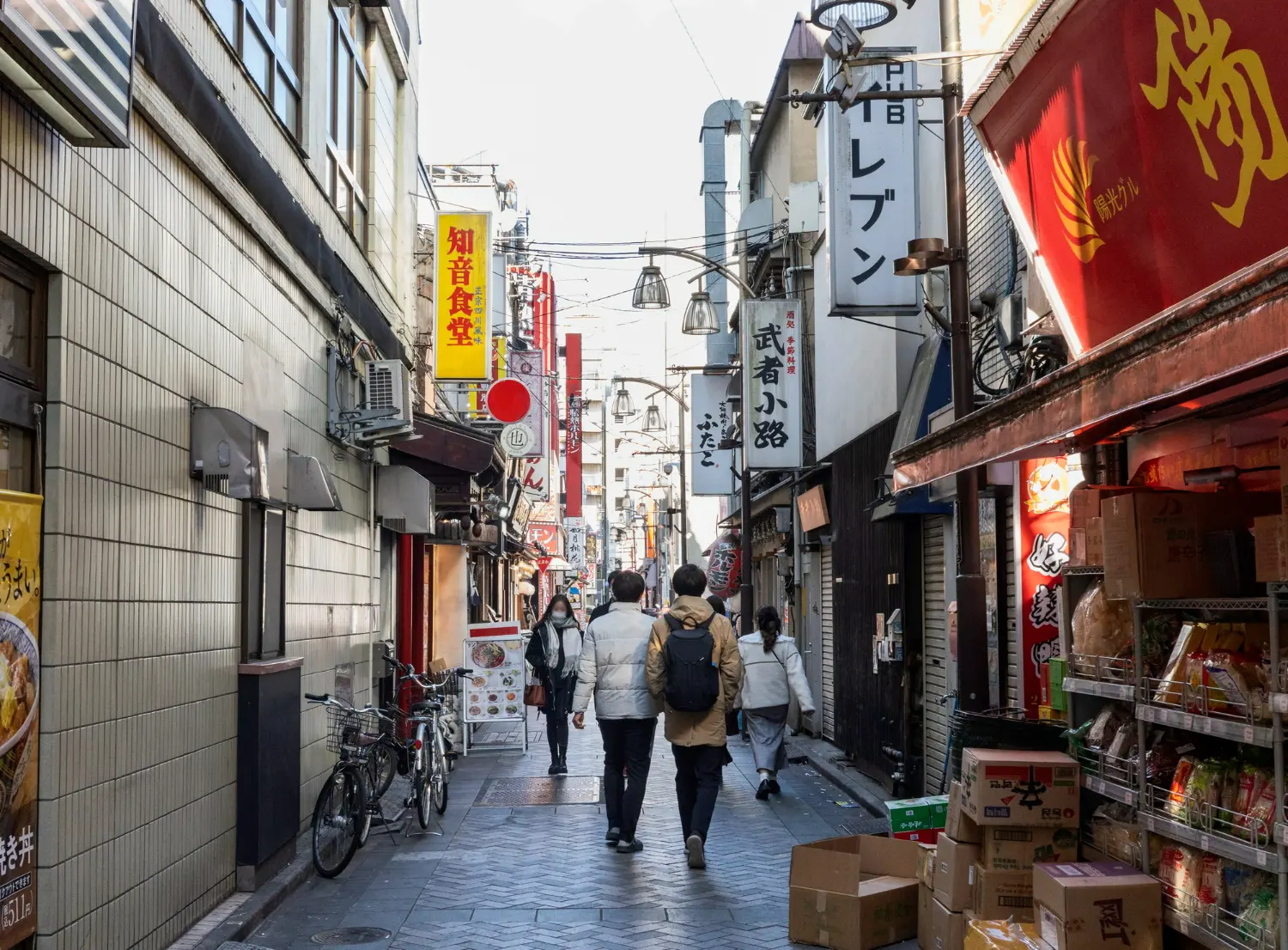 A narrow outdoor alleyway in Japan lined with small restaurants and shops. Signs in Japanese hang above the street, bicycles are parked against the wall, and a few people walk down the path in coats on a sunny day