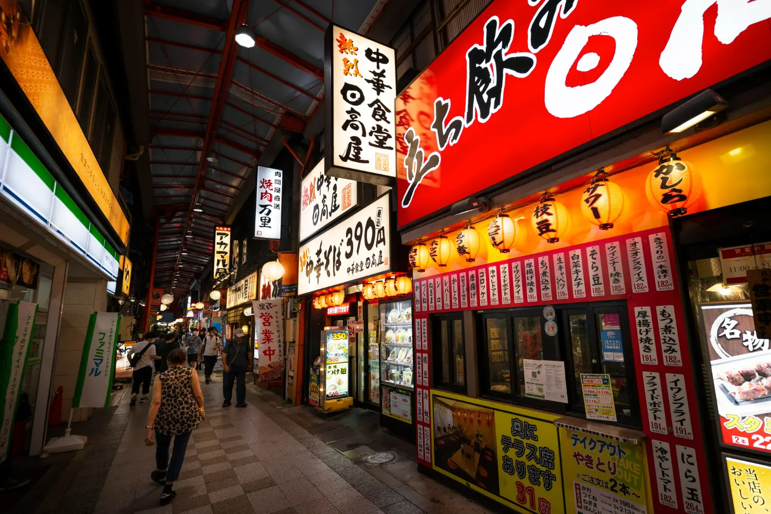 An indoor Japanese shopping arcade with colorful signs, restaurants, and food displays. People casually stroll through the covered walkway with bright lights illuminating the market stalls and eateries