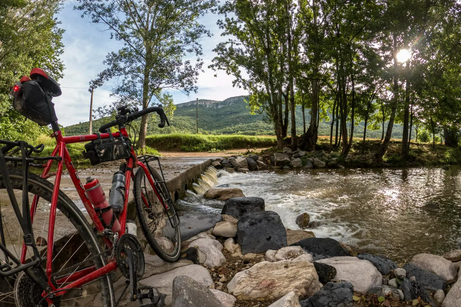 A loaded red touring bike resting beside a shallow stream crossing with tree-lined mountains in the background during a Wales bikepacking trip