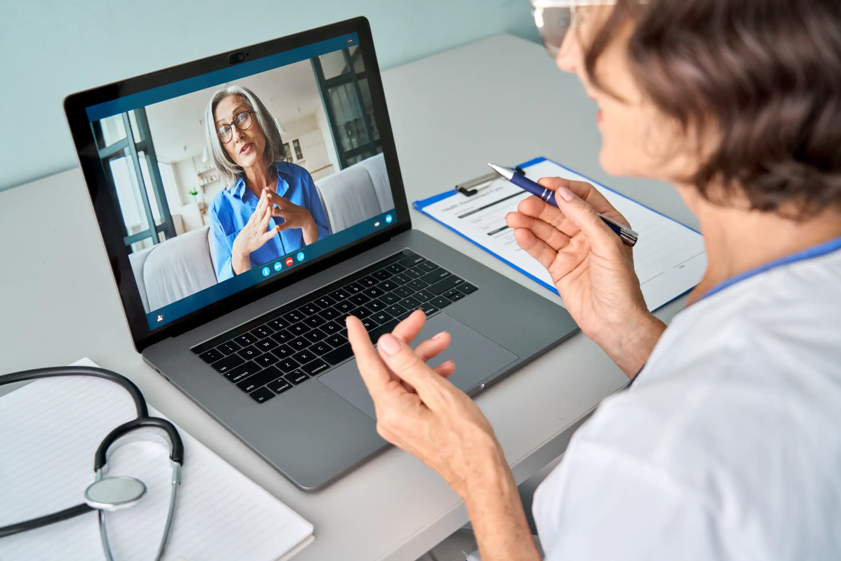 Female doctor therapist consulting older senior patient via virtual video call visit using laptop computer. Digital online healthcare, distance telemedicine. Telehealth videocall. Over shoulder view