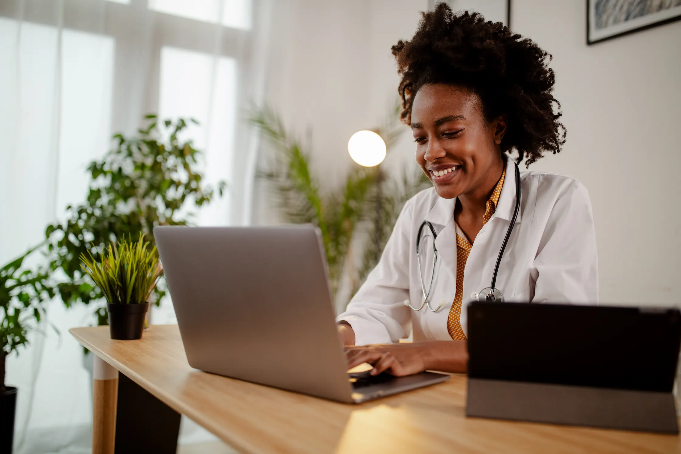 Successful young Afro American female doctor in white medical uniform and stethoscope busy working on laptop in modern hospital, smiling woman nurse or GP typing on computer consult patient online.
