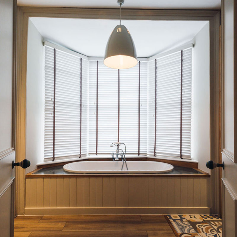 Sunken bathtub with wooden paneling below a large window covered by white blinds and a hanging ceiling light above.