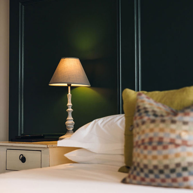 Lit table lamp on white nightstand beside bed with white sheets and green and multicolored pillows.