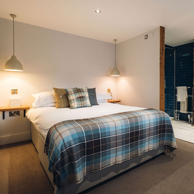Modern bedroom with a large bed featuring white linens, plaid pillows, and a matching plaid blanket, flanked by hanging pendant lights and wooden bedside shelves.