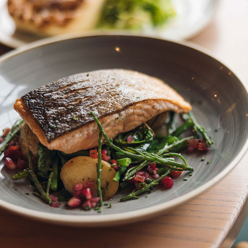 Seared salmon fillet with crispy skin served on a bed of sautéed greens, potatoes, and garnished with pomegranate seeds in a gray bowl.