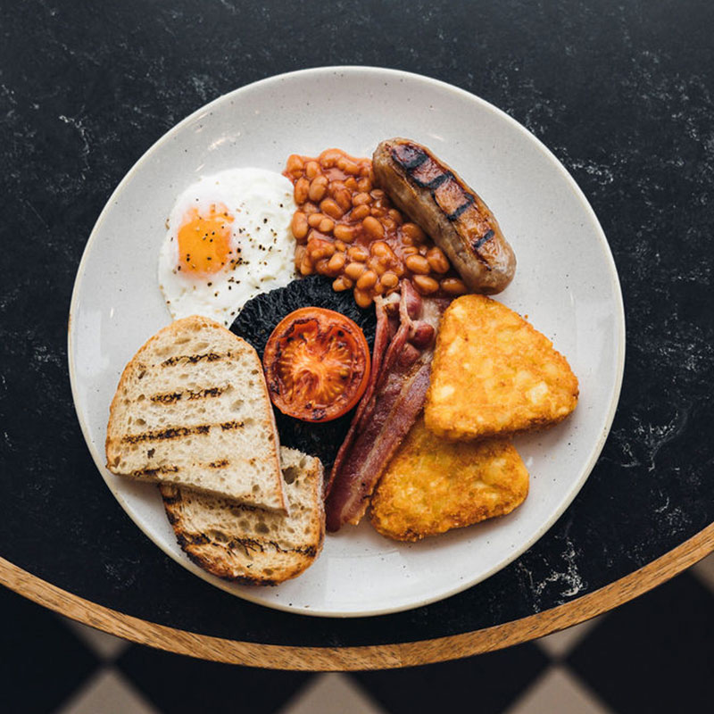Full English breakfast plate with fried egg, baked beans, grilled sausage, bacon, grilled tomato, mushrooms, hash browns, and toasted bread.